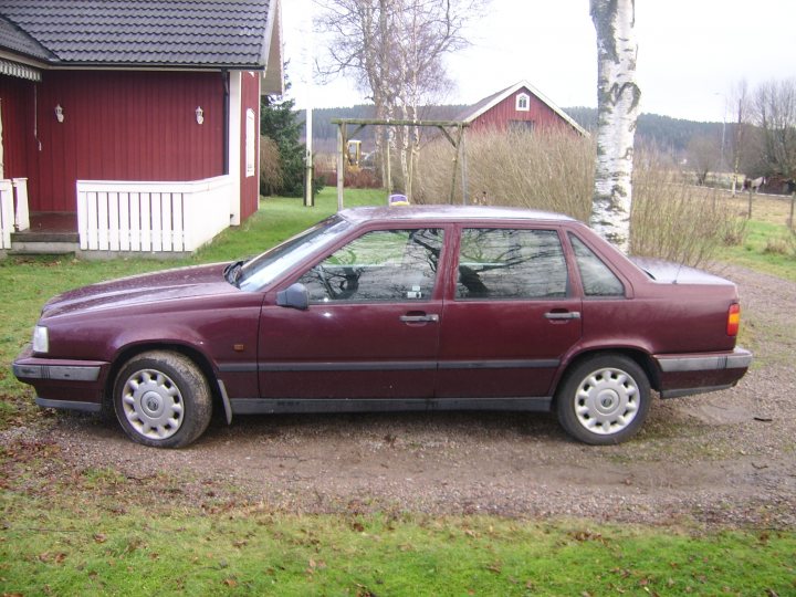 Volvo Owners Pistonheads Aston - The image depicts a maroon-colored sedan parked on gravel in front of a red barn. The car has a sophisticated design with a prominent grille and intricate wheel covers. The surrounding environment features a mix of grass and trees, suggesting a rural or semi-rural setting. There's a wooden structure with a matching red roof to the left of the photo, while further in the background, you can see additional barns and a green field. The scene appears serene and untouched, with no visible signs of recent activity.