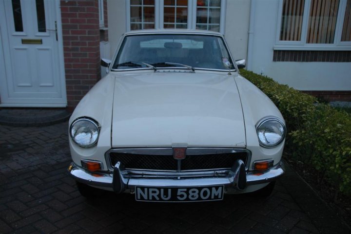 Pistonheads Hurt - The image features a classic white sports car parked in front of a white house. The car has a black monogram on its side, and it is under a car port. In the foreground, there's a brick driveway leading to the house, and a white door can be seen to the left of the car. The setting gives off a nostalgic and elegant vibe, reminiscent of the 1960s or 1970s.