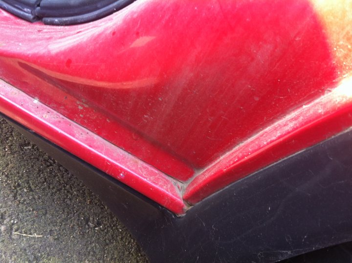 Pictures of my "NEW" SD1 Vitesse - Page 1 - Rover - PistonHeads - The image features a close-up of a vehicle's front bumper, which is prominently painted in a vibrant red color. The red paint has begun to peel and chip away, revealing the black material beneath, which appears to be just paint covered by plastic cladding. The bumper has a slight indentation on the lower left side, which adds to its weathered appearance. Bits of debris, perhaps from the road, can be observed on the bumper's surface. The photo provides a clear view of the texture and condition of the vehicle's bumper.