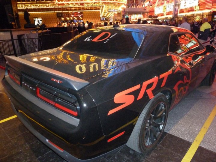 Who else out there has an order in for a Challenger Hellcat - Page 1 - Yank Motors - PistonHeads - The image showcases a sleek black car on display at a convention or show. The car has a distinctive, glossy appearance and features red and orange text on its side and back, which appears to be the name "STRT" repeated. The car is parked in a space with a yellow marking, indicating it's part of a set display. The setting suggests a crowded indoor event with banners and signs in the background, contributing to the ambiance of a convention or exhibition.