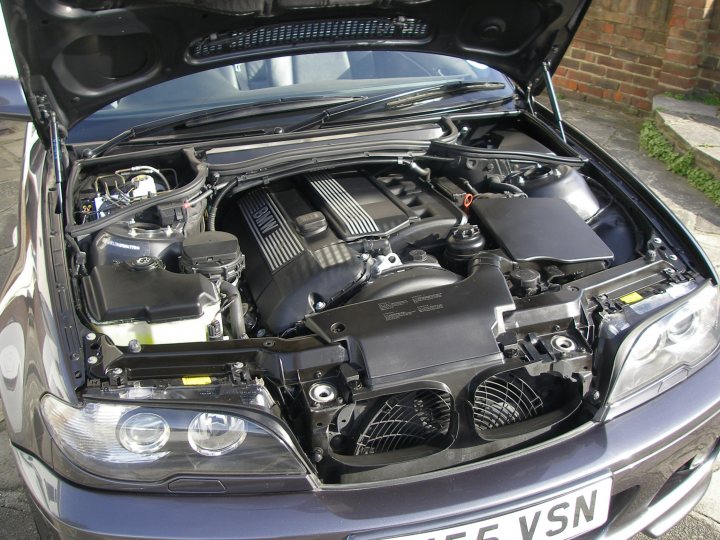 Pistonheads - The image shows the front end of a vehicle parked on a sidewalk. The bonnet (lifted hood) is opened, revealing the engine with black components and wiringrunning through it. The car's lights are yet to be installed. The engine space appears to be dirty or contains dust and small debris. The vehicle is parked closely to a brick wall in a city setting.