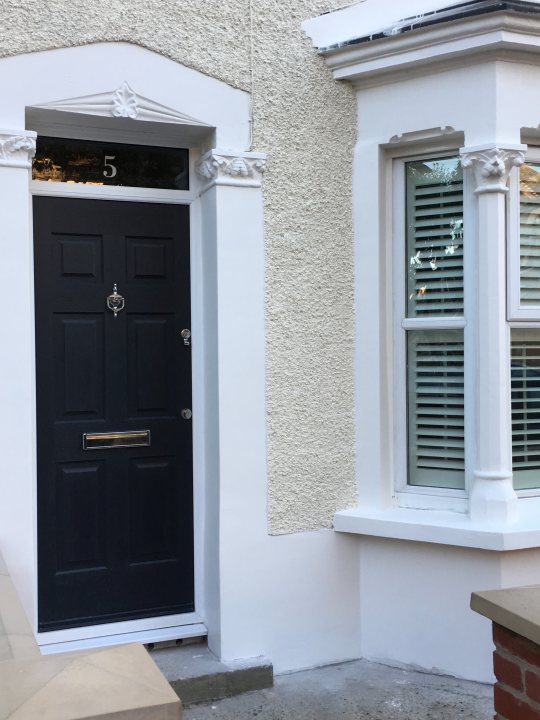 Composite Doors - HELP! - Page 1 - Homes, Gardens and DIY - PistonHeads - This image features a white two-story dwelling with architectural details. The main entrance door is black, probably painted, with a small window above it and a number '5' visible above it. The door also comes equipped with a mail slot and a peephole visible at eye level. There's another window on the right side of the house, and the windows seem to be shuttered or closed curtains. Additionally, a decorative column or cement feature is present, positioned on the ground level on the right side. The house appears to be corner plaster construction with a contrasted light stone facade.