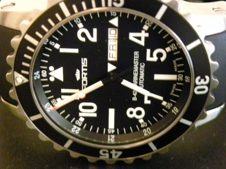 Pistonheads Collection - The image features a close-up of a round dive watch. The watch face is black and orange, with the numbers 12, 6, and 3 prominently displayed. The dial includes multiple smaller indicators like goal posts, directional arrows, and a compass rose, along with hour markers. The watch has a silver metal band and a stainless steel case, conveying a sense of robustness and precision.