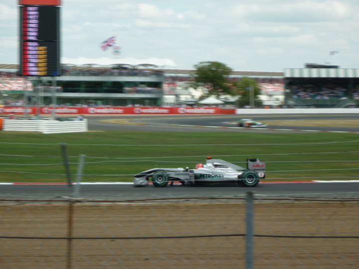 The Official Silverstone GP thread 2014 ****SPOILERS**** - Page 1 - Formula 1 - PistonHeads - The image captures an intense moment at a motor race. A car in the blue and green color scheme is dominating the front of the scene, likely leading the lap. The setting appears to be a large racing circuit with stadium-style seating visible in the background. The photo is taken during the day under clear skies, and the focus is solely on the race track and its competitor. The image conveys the speed, precision, and competitiveness of the race.