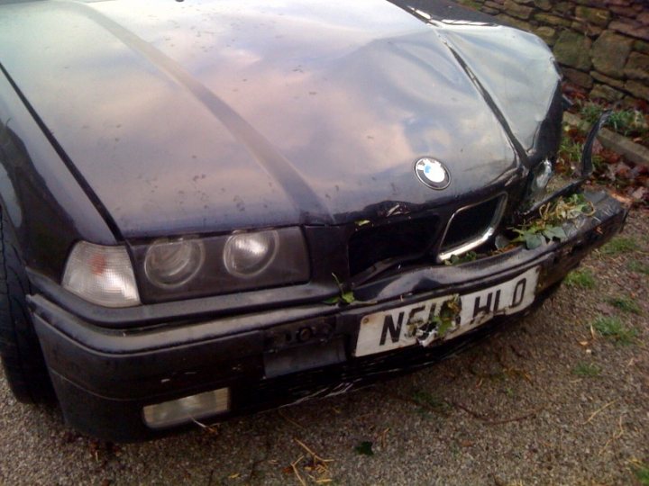 Pistonheads Daily Peugeot Snotter Free - The image shows a black BMW car with a severely damaged front end. The hood is crumpled, the bumper is bent, and the grille is shattered. The car has left its intended use on the road and now rests on a patch of grass, accompanied by a wall in the background, adding to the sense of abandonment. The overall scene suggests some form of accident or abuse has taken place, with the damage fully on display.