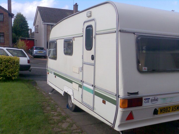 My caravan history - Page 1 - Tents, Caravans & Motorhomes - PistonHeads - The image shows a caravan parked on a grassy area in a residential area. The caravan is primarily white with horizontal stripes in green and olive colors. The rear of the caravan has a license plate, a holder for a gas can, and a small red triangle. The caravan's roof camps, indicating it is designed for camping or mobile living. The grass appears well-maintained, and there are no people visible in the image. In the background, there is a house with a red door, and a car is parked in front of a neighboring house. The sky is overcast, suggesting a cloudy day.