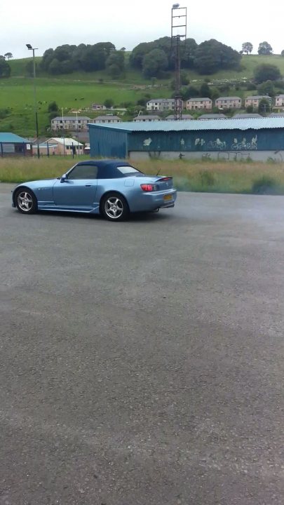 my honda s - Page 1 - Readers' Cars - PistonHeads - The image captures a scene of a blue sports car parked on a concrete surface of a dark gray color. The car is facing towards the left side of the image. The background provides a contrasting view of a green hill or slope with houses scattered along its length, suggesting a residential area. To the right, there appears to be a gate, indicating a boundary or entrance to a property or an avenue. The weather seems overcast, with no visible clouds. The setting appears to be during the daytime.