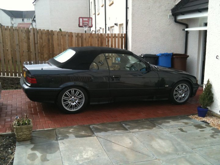 Our Cars - Page 40 - Scotland - PistonHeads - The image shows a black convertible sports car parked on a red brick driveway in front of a house. The vehicle has a convertible top up and features multi-spoke wheels and a sleek design. In the background, there's a wooden fence and a white building beyond it. A blue bucket sits on the sidewalk next to the car, suggesting activity or maintenance. The sky is emphasized in the photo, with a clear hint of blue, which complements the overall color palette.