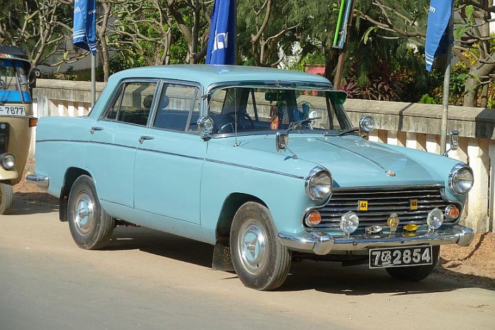 What is this car spotted in Sri Lanka - Page 1 - Classic Cars and Yesterday's Heroes - PistonHeads - The image showcases a vintage E-type (7523854) Jaguar car painted in a shade of light blue, parked on a residential street. The car's license plate gives away its origin and model year. The rearview mirrors shine in the sunlight illuminating the scene. The surrounding environment is blurred, but one can make out the tranquil setting with trees and a roadside fence. A few flags can also be spotted in the scene, adding a festive touch to the backdrop.