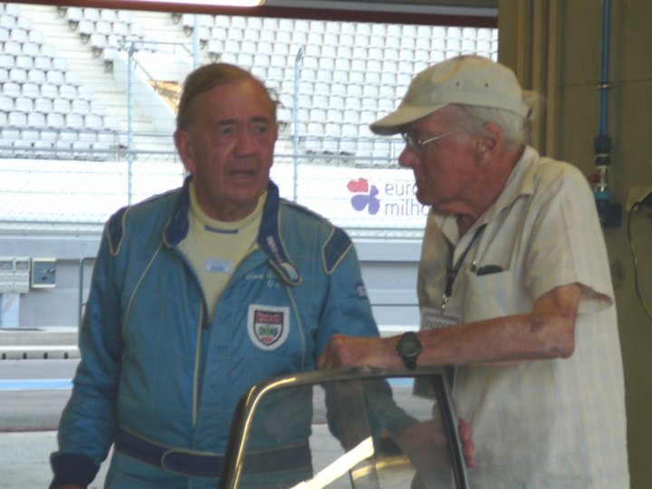 Founder Rip Ralph Broad Broadspeed Pistonheads - The image portrays an older man and a younger man engaged in conversation inside what appears to be a metal, semicircular enclosure with a racetrack background. The older man is wearing glasses and a baseball cap, while the younger man, dressed in a blue racing suit, has his own baseball cap and his back to the camera. Both are looking towards the right side of the image. The casual confrontation between the two men, along with the distinctive attire of the younger man, suggests a possible event or gathering related to motorsports.