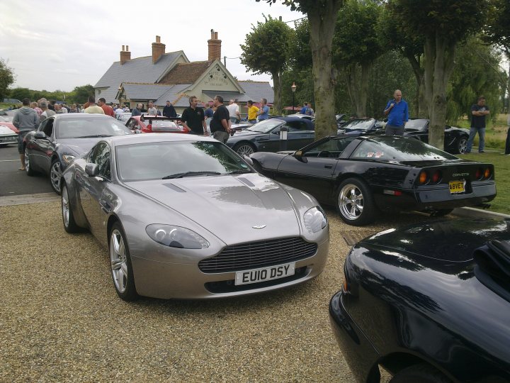 Pistonheads - This is a daytime photo showing a line of modern sports cars parked on gravel, likely on a sunny day given the shadows. Among the cars, there are two prominent silver cars, one of which may be an Aston Martin judging by the distinctive grille, suggesting they are in a small parking area or on display at an outdoor event. The people next to the cars are casual and appear to be attendees or participants at the same event. The vehicles, with their shiny exteriors and vibrant colors, are the primary focus of the image.