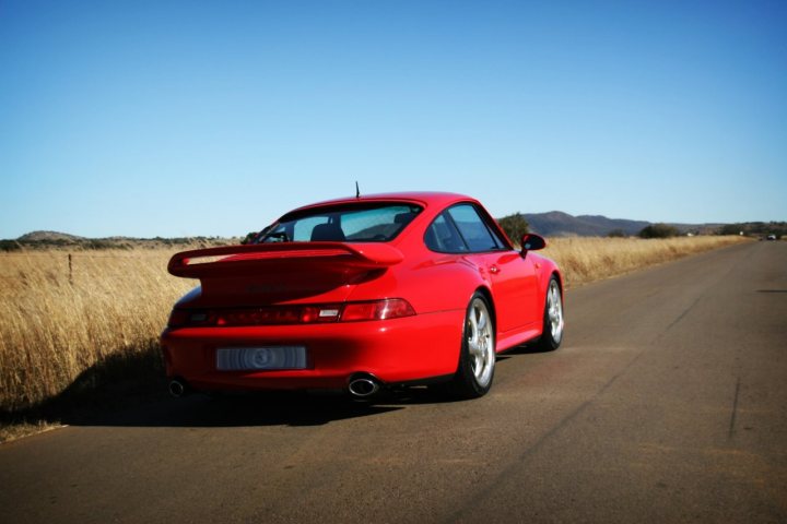 Some 993's - Page 1 - Porsche General - PistonHeads - The image depicts a vibrant red sports car parked on the side of a desert road. The car's bodywork is beautifully smooth, reflecting the sunlight. The car is angled towards the viewer, showcasing its design features, such as the large air intake vents and rims. The backdrop of a wide-open desert and a clear blue sky gives the scene a sense of space and freedom.