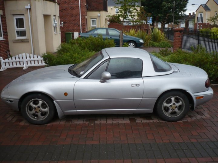 Pistonheads Booked Bodyshop - The image shows a small two-door sports car parked on a residential street. It has a sleek, silver body with a convertible top that is currently up. The car is parked on a driveway of a brick home, and there is another vehicle parked next to it. It is a clear day, and the surroundings of the car include greenery and other residential structures.