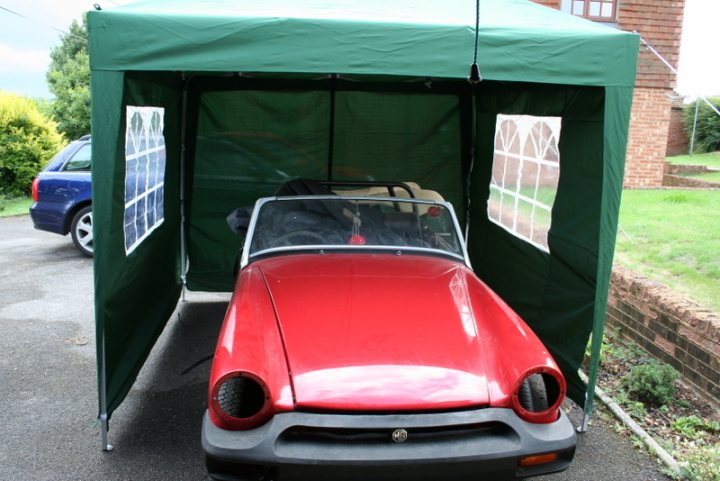 MG Midget - Just get it finished !! - Page 1 - MG - PistonHeads - The image showcases a red MG convertible car. It is prominently displayed inside a green pop-up tent, which is placed on what appears to be a residential driveway. In the background, there are two vehicles: a blue station wagon and another compact car. The overall setting suggests an outdoor event or gathering, possibly for the classic car.