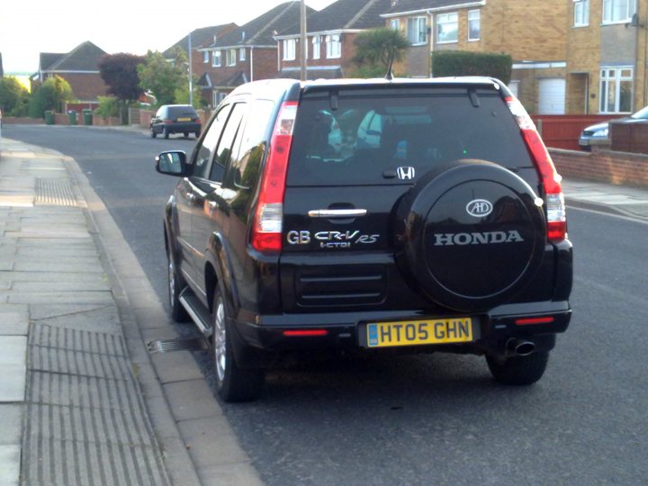 Misbadging Pistonheads - The image features a dark-colored car parked on the side of a street in a residential area. Behind the car, we can see a typical suburban scene with houses, a fence, and another vehicle parked further down the street. The car is labeled with the letter "H" and the model "GS CVES" suggesting it's a model by Honda. The license plate is visible on the back of the car and reads "HT05 GHN." The vehicle appears to be in good condition and is stationary, as indicated by the absence of any motion blur.