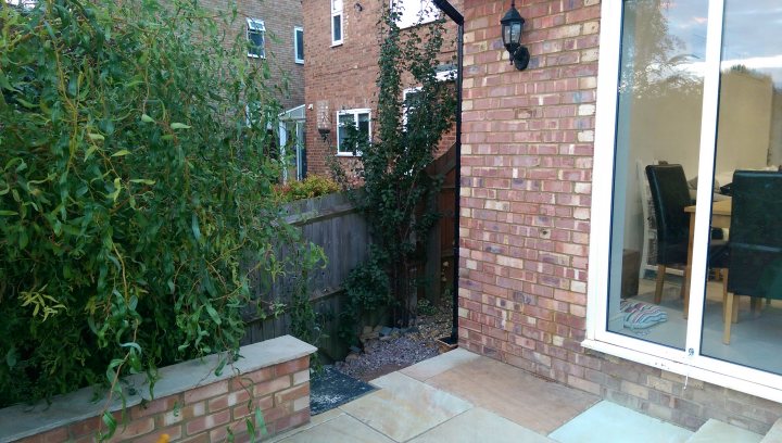 Planning permission for a patio - Page 1 - Homes, Gardens and DIY - PistonHeads - The image presents a well-manicured front yard of a house. A red brick wall defines the front yard, with a decorative white fence on top. Beside the entrance is a black wrought iron light fixture, adding a touch of elegance. A glass door with a white border reveals a serene interior with a dining table and chairs visible through the window. A black leather sofa is situated next to a window that lets in soft natural light. The overall scene is a harmonious blend of outdoor and indoor elements, creating an inviting atmosphere.