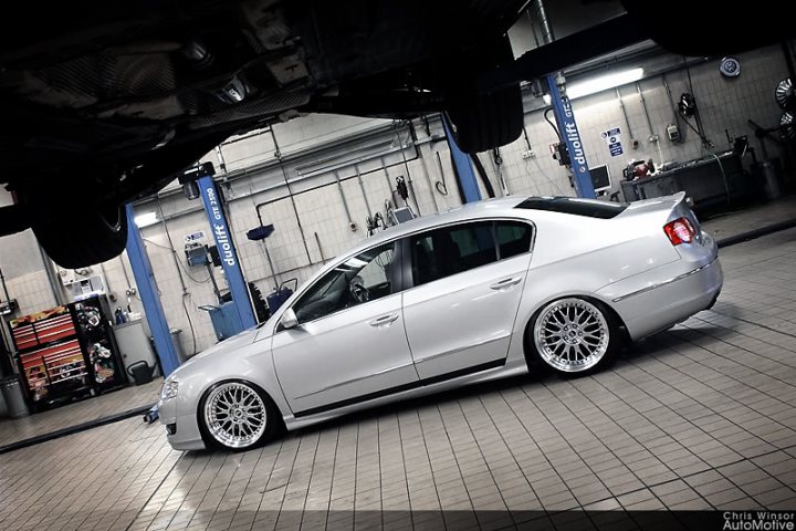 Pistonheads Dub Scene - The image presents a scene from a workshop, where a silver sedan, equipped with a wide-body kit that adds to its dynamic appearance, is parked on a tiled floor. The vehicle features a large, aftermarket spoiler on its trunk and a distinctive design element known as "wolf" wheels. The surroundings include various equipment and tools associated with automotive servicing.