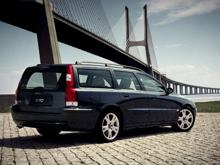 2006 V70 rear fog lights - Page 1 - Volvo - PistonHeads - The image showcases a dark blue station wagon parked on a cobblestone area adjacent to a body of water. In the background, there is a large suspension bridge with cables stretching across the bridge. The sky in the background features a few scattered clouds. The car's reflections can be seen on the glossy surface of the water, and the time on the car's clock reads "1:20 PM".