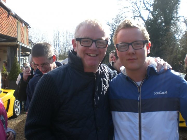 Pistonheads Bragging Evanss Kid Chris - In the image, there are two men standing close together, smiling broadly at the camera. The man on the left is taller with dark hair and glasses, while the man on the right is slightly shorter with light brown hair, also wearing glasses. They are dressed casually, with the man on the right sporting a blue and white striped jacket. In the background, there are other people and a yellow vehicle, suggesting they might be at an event or gathering.