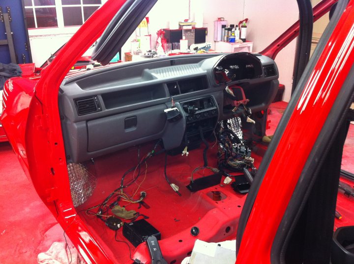 Pistonheads - The image showcases the interior of a red car, specifically focusing on the open dash area. The car seems to be on a lift, suggesting that it is in the process of being repaired or maintained, as evidenced by the visible wires and parts scattered around. Various tools and components are spread out across the floor, with bottles and other smaller items scattered around the car. The red car's door is open, providing a clear view into its interior. The image conveys a sense of activity and maintenance, with the car seemingly in the process of repair or improvement.