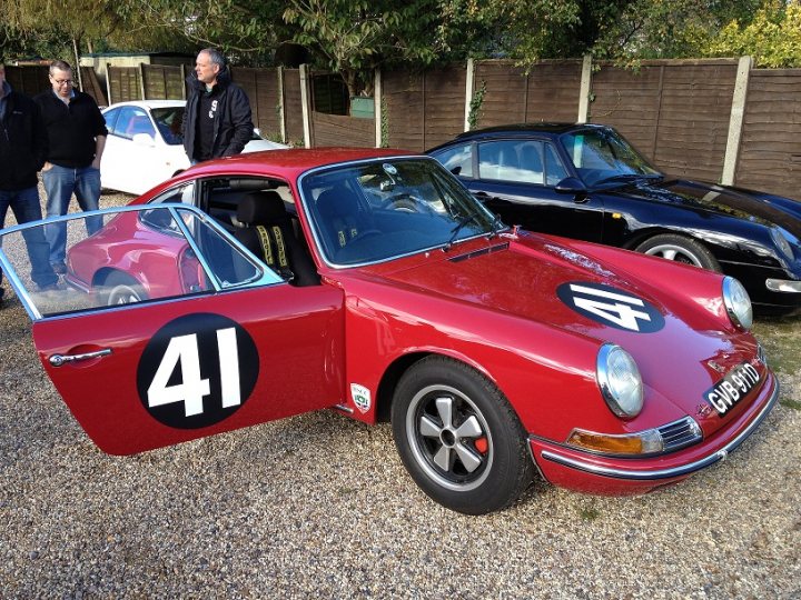 A red car is parked in a parking lot - Pistonheads - The image showcases a gathering of vintage racing cars, all parked neatly on a gravel lot. Each car boasts a unique color and design. In the foreground, a striking red classic car commands attention, its door open to reveal a white driver's seat. The white car parked next to it has its door closed, offering a different perspective. Both cars are secured to a wooden post, ensuring their stability on the gravel. The setting is serene, with a backdrop of a lush garden under a clear blue sky.