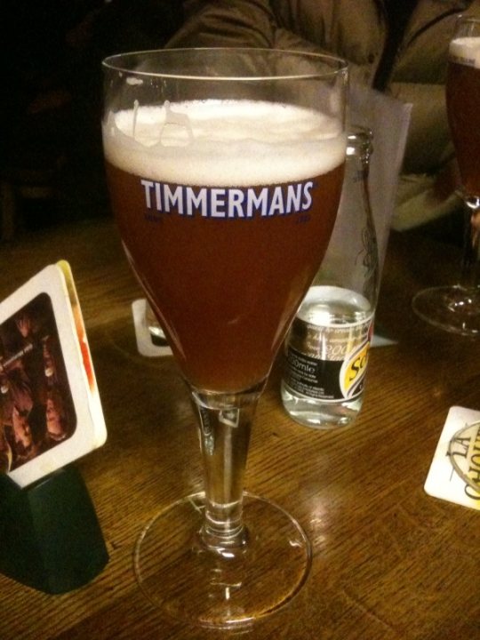 Interesting Lager Beer Pistonheads - The image showcases a tall glass prominently placed on a wooden table. The glass is filled with a frothy beverage, emitting a white foam at the top, suggesting it's a craft beer. On the left side of the image, partially out of focus, there's a smaller glass next to the main glass, suggesting there might be another beer being served. The wooden table upon which the glasses rest has a beer coaster on it, adding to the cozy ambiance of the scene. The label on the glass reads "TIMERMANS", indicating the brand of the beer. The overall atmosphere of the image suggests a relaxed, casual social setting, possibly at a pub or a brewery.