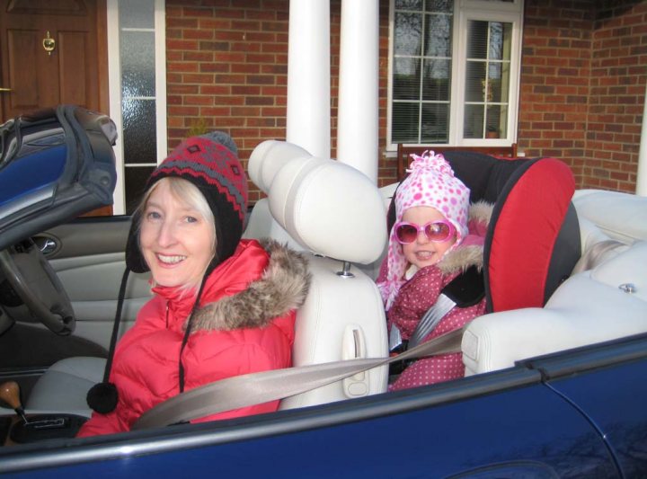 In the image, a woman aboard a blue convertible car is wearing a red beanie and a pink puffer coat, her smile radiating warmth. A young child, adorned with a pink hat, pink sunglasses, and a black jacket, is seated beside her, sharing cheerful expressions. The car's interior is visible, featuring white leather seats, and the car is parked outside a brick house, with a row of windows reflecting the residential setting.