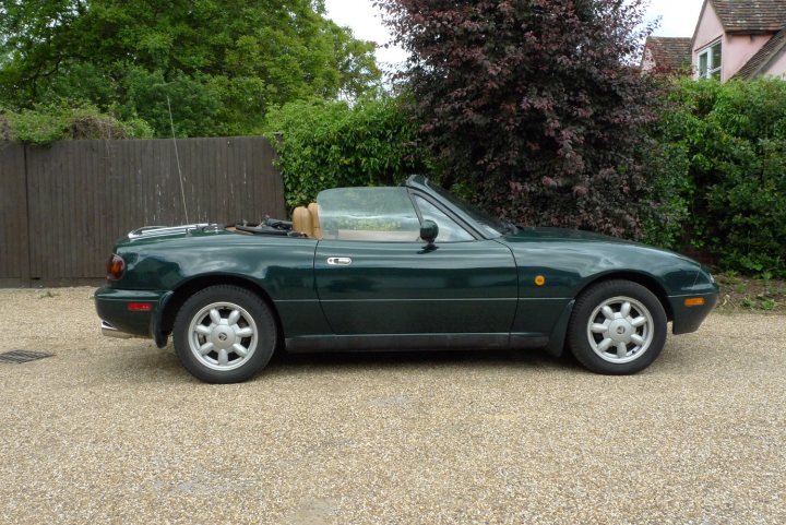 Mazda Pistonheads - In this image, there is a stylish convertible car parked on a gravel driveway. The car, painted in a deep green color, is the main focus of the image. It's a two-door model with the roof down, revealing two round seats indicating it's designed for two passengers. The car is positioned on the right side of the image, with the driveway extending down the middle. The setting appears to be suburban, with trees and a house visible in the background, suggesting a residential area.