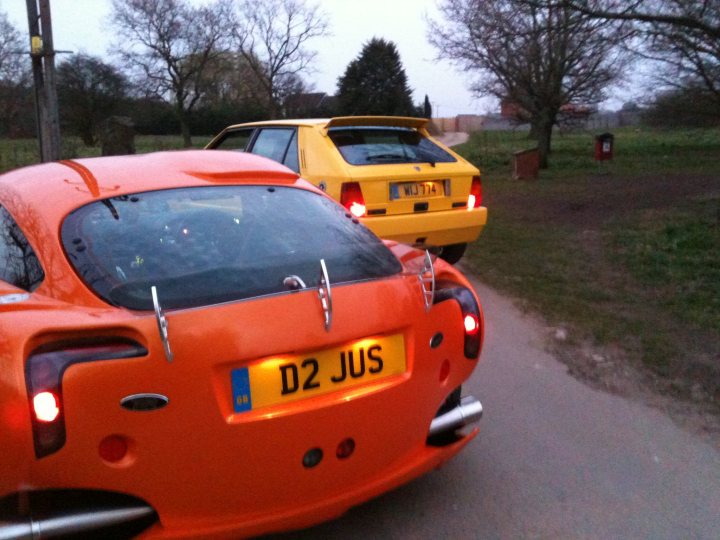 Pistonheads Seconds Endulge - The image shows a scene of two cars on a road. The cars appear to be of a similar make and model, though one is a bright orange and the other is a soft yellow, distinguishable in the lighting. The first car has glowing red taillights and is approaching, while the second car has braking lights illuminated, suggesting it may be following a slower car or stopping. The backdrop is a darkened evening sky with trees, and the road seems relatively empty aside from these two cars.