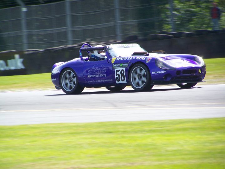 Oulton Park Tvr Pistonheads Sat Challenge October - The image captures a dynamic scene on a race track. A sleek purple sports car, bearing the number 58, is in the midst of a high-speed turn. The car's vibrant color stands out against the green of the track and the blur indicating its speed. In the background, there's a person and what appears to be a building or a fence, possibly part of the racing infrastructure. The photo captures the car's front, emphasizing the driver's focus and the power that this machine is unleashing.