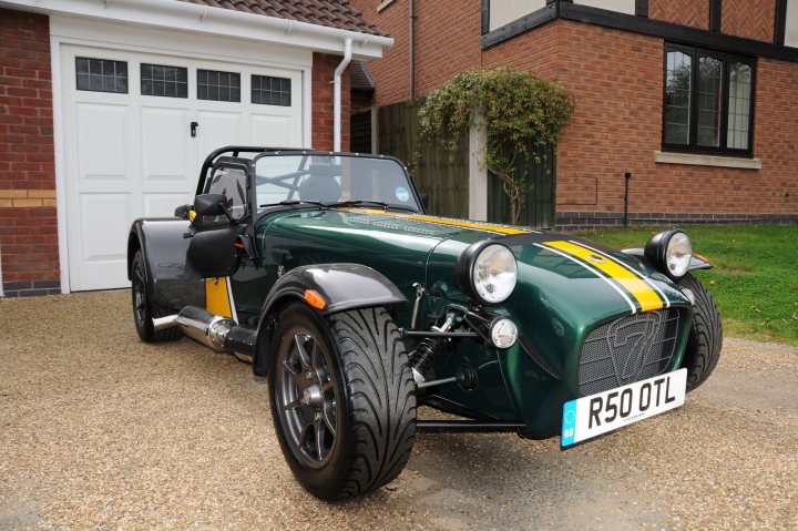 caterham racing colour code? - Page 1 - Caterham - PistonHeads - The image showcases a striking black and green sports car parked in front of a brick house. The car is a rare sight, equipped with a roll bar and what appears to be a batmobile-esque design, featuring a wide hood and a sleek, low-to-the-ground profile. Its color scheme, with contrasting accents, reflects a unique blend of aesthetics and performance. The house in the background has a quaint charm with a brick facade and a neatly trimmed garden, providing a serene backdrop to the car's imposing presence.