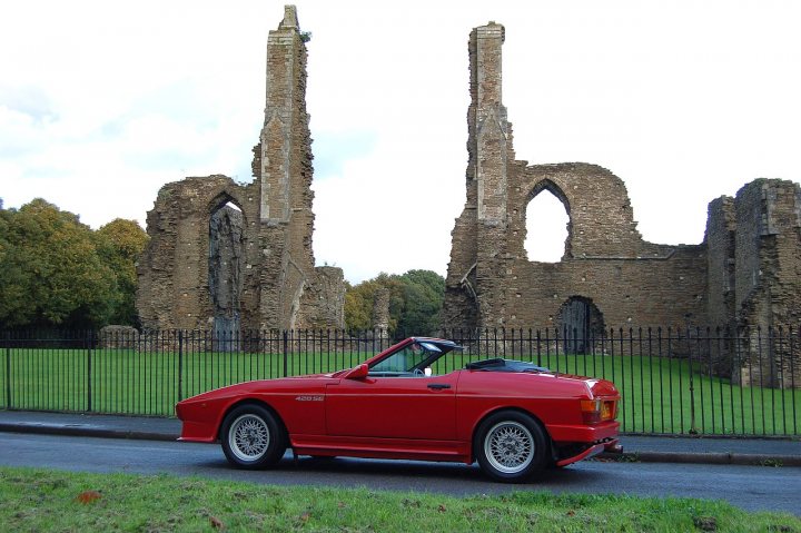 Places to take good photos... - Page 1 - South Wales - PistonHeads - The image depicts a red sports car parked on a paved roadway. The car's design is reminiscent of classic models. Behind the car stands an imposing structure, which is a large, ruined stone building with crumbling walls and two prominent stone arches. The setting seems to be in the countryside, with grassy lawns and trees surrounding the building. The sky is partly cloudy, suggesting a day with some variation in weather conditions.