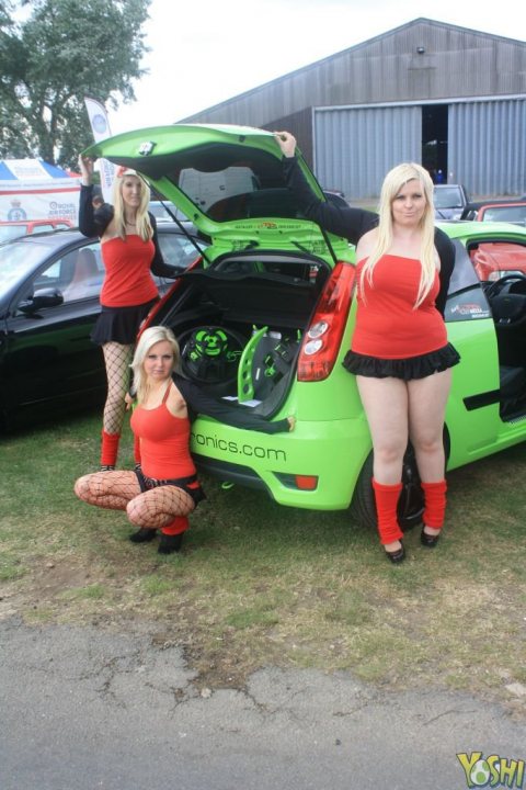 Grid Girls/Pit Babes (Vol 3) - The image captures a lively scene in a parking lot dominated by a striking green VW GTI hatchback. Two women, one on either side of the car, stand out with their revealing outfits. The woman on the right pairs a vibrant red blouse with a black skirt and red fishnet stockings, while her companion to the left sports a matching red blouse, a black skirt, and black fishnet stockings. Their confident poses encapsulate a sense of flair and youthful energy. In the background, other parked cars can be seen, creating a diverse tableau of colors and shapes.