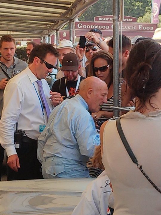 Post your Pictures here: Festival of speed 2013 - Page 1 - Goodwood Events - PistonHeads - The image captures a buzzing scene with a group of people gathered around a man in a white shirt with sunglasses. The focus of the scene is clearly on the man, who is standing under a tarp and appears to be signing autographs. The man is surrounded by a crowd of people, some of whom are taking pictures while others are handing him things. The tarp suggests it might be an outdoor event, possibly related to the gentleman's interests or profession. The atmosphere seems lively and engaged, indicative of a public activity or appearance.