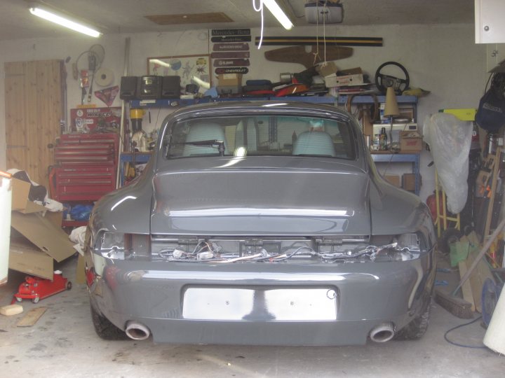 A car that is sitting in the street - Pistonheads - The image shows a dark gray or silver, two-door vehicle, likely a convertible or coupe, in the midst of being worked on or is undergoing restoration in a garage or workshop. The car is positioned against the back wall, which is cluttered with various technical tools, shelves holding miscellaneous items and equipment, and other vehicles. The setting is well-lit by fluorescent lights and is characterized by an open door and an array of mechanical parts and tools scattered around. The car appears to be missing a rear window or window frame and may require bodywork or repairs before it's road-worthy again.