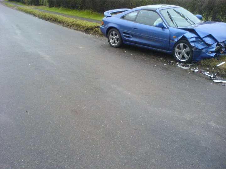 She's a write off. - Page 2 - Jap Chat - PistonHeads - The image presents a blue car that has been involved in a severe accident. The car is parked on the side of a street with grassy areas in the background. One side of the car is visibly damaged, displaying a crumpled front end and a bent wheel. It's important to note that the image depicts a wreckage and the occupants of the car are not visible.