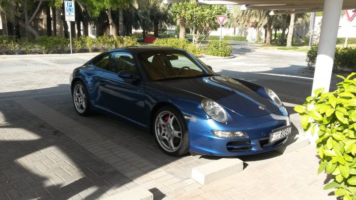 New Wheels - Page 1 - Middle East - PistonHeads - The image features a **blue Porsche sports car** parked in a **bricked parking space**. The car's sleek design and distinctive badge are clearly visible. The setting appears to be an outdoor parking lot, with a **building** and a row of trees in the background. There are parking signs visible, including a **blue and white sign** in the background. The parking lot has a **paving stone surface**, adding to the urban feel of the scene. The time of day seems to be during the day, as there is bright sunlight illuminating the scene.