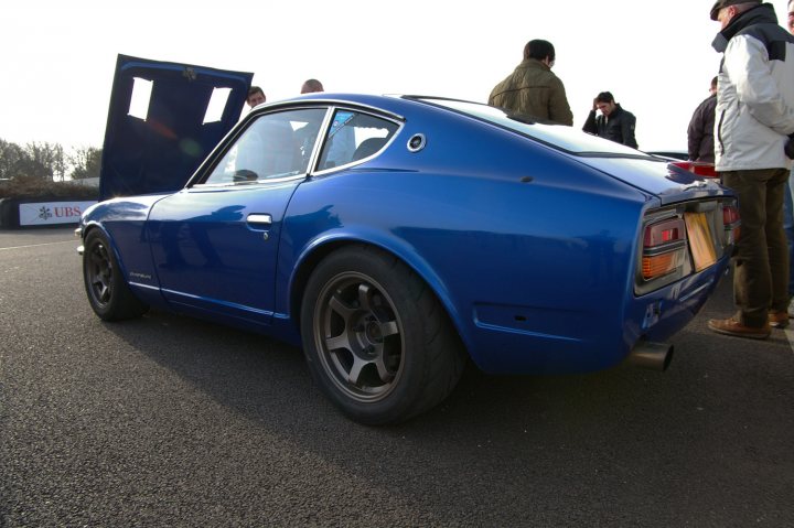 Goodwood Breakfast Club - Sushi Sunday - photos - Page 1 - Goodwood Events - PistonHeads - The image captures a scene with a vibrant blue sports car as the main focus. The car is positioned on what appears to be a road, with its hood open, revealing the undercarriage. The environment suggests an outdoor setting, possibly near a car show or auto event. There are several people visible in the background, walking away from the car, indicating that they may have been inspecting it or admiring its design. The overall atmosphere is dynamic and charged with anticipation for the start of the event or journey.