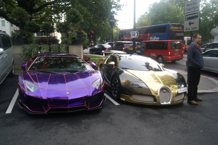 A bunch of cars that are parked in a lot - Pistonheads - The image shows a street scene with several vehicles, the main focus being on two luxury cars, one purple with designs around its hood and the other gold with a similar design on its hood. The cars are parked in a complex giving access to various locations and services, including what seems to be a hotel and a parking structure. Other cars and buses are visible in the background, behind a tree and amidst various plants and a fountain, signifying a city or urban environment. There's a mix of both stationary and moving vehicles, suggesting normal traffic on a clear day.