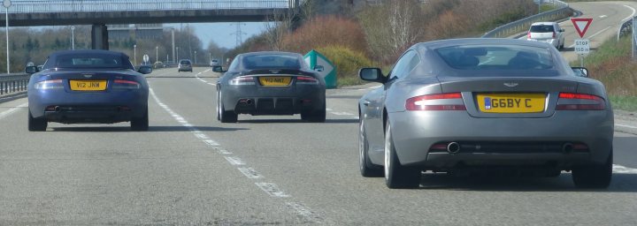 How about an Aston photo thread! - Page 68 - Aston Martin - PistonHeads - The image shows a gray car on a highway, described as "losing his lunch." There are two dark-colored vehicles behind it. There are distinct yellow license plates and white brake lights. This suggests a scene possibly from the early to mid-20th century, as these style of cars and license plates are common in that era.