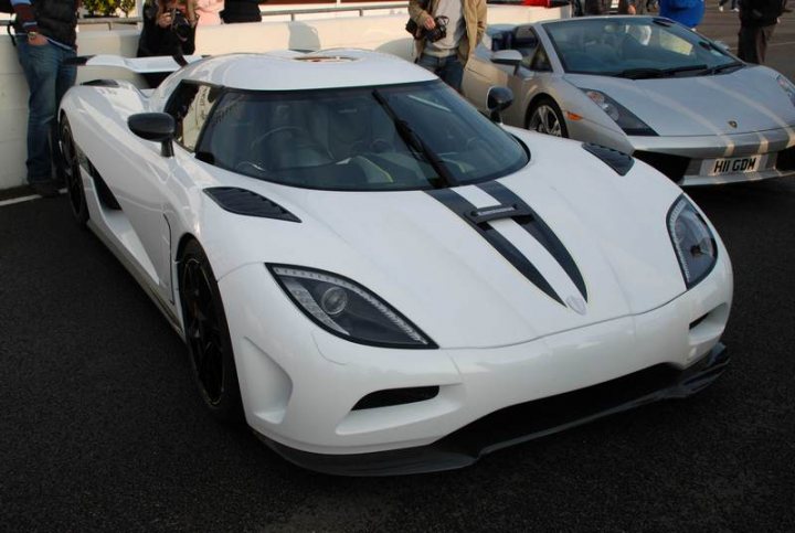 Sunday Supercar Pistonheads - The image showcases a white sports car parked on a road. The car is sleek and modern, characterized by its long hood, short rear deck, and distinctive LED headlights. In the background, there are people and additional cars visible, though they are not the primary focus. The setting appears to be an outdoor event or gathering where the sports car is on display.