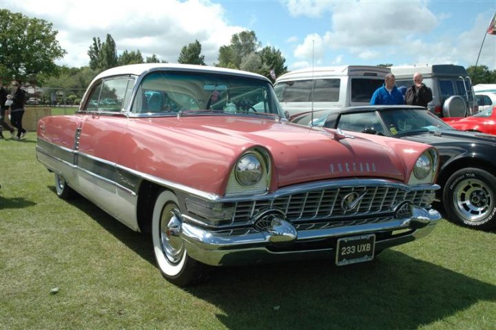 Fins and Chrome - Page 1 - North East - PistonHeads - The image depicts an outdoor scene, with some emphasis on several vintage cars. There is a classic car in the foreground, painted in a shade of pink that gives it a vibrant and nostalgic appearance. This car features a silver color scheme on the lower section, including the hubcaps and a metal trim.

Additional cars are present in the background; these include a black car and a white RV-style trailer. On the far right, another vintage car is situated at an angle, adding variety to the composition.

The setting appears to be a grassy area that might be part of a campground or similar outdoor location. A few people can be seen in the distance near the vehicles, possibly enjoying some kind of gathering or event. The grass and trees suggest a pleasant day with clear skies. The style of the car in the foreground, along with the rest of the scene, evokes a sense of leisure and appreciation for old-school automobiles.