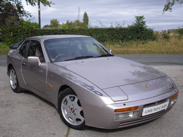 944 Silverrose - Page 1 - Front Engined Porsches - PistonHeads - This image depicts an illuminated, sleek silver Porsche car parked outside with a slightly upward angle, giving a view of its front and side. The license plate is visible, reading "GMUND CARS," and on the side of the car, there's a script that says "GE-GE". The surroundings show grass and bushes, suggesting a casual, outdoor setting. The car's design features smooth lines and standard side mirrors.