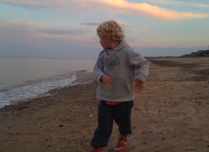 Lowestoft - Page 2 - East Anglia - PistonHeads - This image captures a heartwarming moment at a beach. A young boy, standing on the sandy beach, is looking out at the vast ocean. He's dressed in loose-fitting blue jeans and a gray sweatshirt, adding a cozy contrast to the natural landscape around him. His curly hair adds a touch of innocence and charm to the scene. It's a beautiful day with a partly cloudy sky, and the sun seems to be setting, casting a warm glow on everything. The beach appears to be empty, suggesting that the boy is there to truly enjoy and appreciate nature by himself.