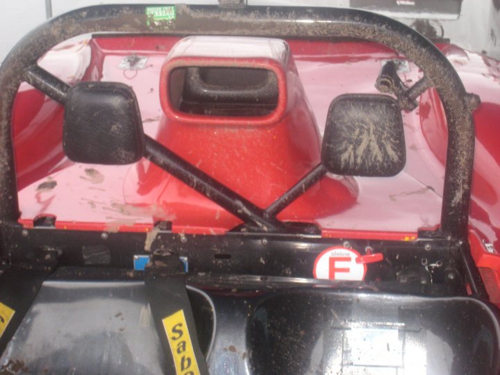 A car is parked on the side of the road - Pistonheads - The image displays a red three-wheeler cart. The front of the cart is predominantly black with space for a seat, indicated by the absence of seat cover. The cart has a smaller backseat visible, still covered with a black seat cover. The microphone stand and the Sygic brand GPS tablet attached to the cart, suggesting it is set up for potential recreational use or for tours. The front of the cart appears a bit dirty and worn, indicating potential outdoor use.