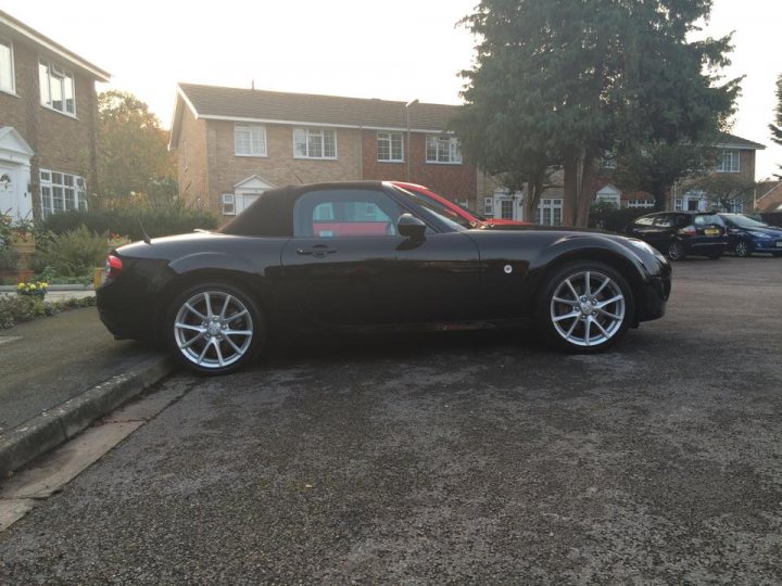 Mk 3 Owners - stiff 6 speed gearbox? - Page 1 - Mazda MX5/Eunos/Miata - PistonHeads - The image is of a two-door black sports car parked on a driveway next to the curb. The car appears to be a convertible or soft-top edition due to the presence of a black fabric top stored in the rear compartment. The vehicle is parked near residential buildings, and there's a paved surface in the foreground that reflects the car and the environment. The sunlight indicates that the photo was taken during the afternoon.