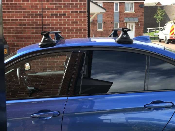 Farad roof bars - wrong way round?  - Page 1 - General Gassing - PistonHeads - This image shows the hood of a blue vehicle with a tinted rear window. On top of the car, there are two ventilation boxes mounted, possibly for a particularly high or fast speed rating. In the background, there's a house and a green lawn, and there's a glimpse of a white construction vehicle. The scene suggests an urban or residential setting.