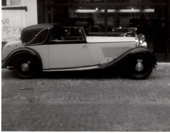 B&W Bentleys.... - Page 1 - Rolls Royce & Bentley - PistonHeads - The image is a black and white photograph of a vintage car parked on a brick street in front of a building. The car has a long body with a coupe design, featuring two small windows behind the front seats and a sunroof above. It has white and black paint, round chrome wheels, and a prominent front grille. The car's front door is partially open, and there is a graffiti-style logo painted on the wall behind the car.