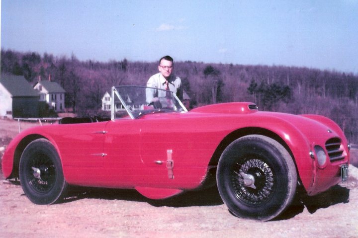 Early TVR Pictures - Page 34 - Classics - PistonHeads - In the image, a man is standing next to a distinctive red Alfa Romeo Spider. The car features a unique silver roll bar, a characteristic design detail of this classic automobile. The vehicle is parked on a dirt road, and the man is captured from a slightly elevated angle, slightly to the left of the car. The backdrop of the image is a blurred landscape of trees, evoking a sense of tranquility and open space.