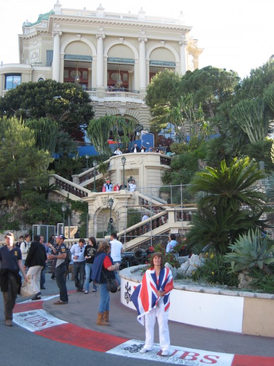 A crowd of people walking down a street - Pistonheads - The image shows a woman standing in the foreground, wearing a dress with a British flag design. Behind her is a large crowd of people, some holding Union Jack flags. To the left is a double-helix structure which could be a monument or exhibit. In the background, a large, ornate building with white architecture is visible, indicating a formal or governmental setting. A portion of a red fence is visible at the bottom of the image. The lighting suggests the photo was taken on a bright, sunny day.